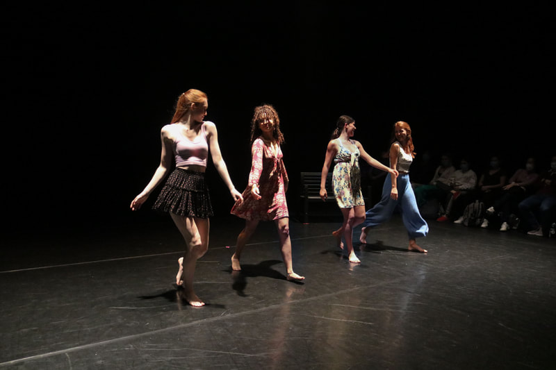 Four dancers, wearing colorful dresses, skirts, and pants, walking on a stage, looking at each other, smiling.