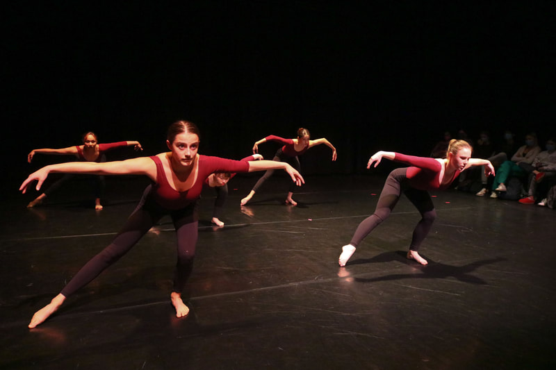 Five dancers in red leotards and black leggings, posing on stage in a tendu with a bent supporting leg and spider arms.