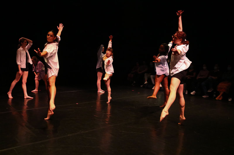 Seven dancers on stage with black shorts and white or blue button-downs. Three front dancers are doing a low kick to the side on relevé. Other dancers are standing.