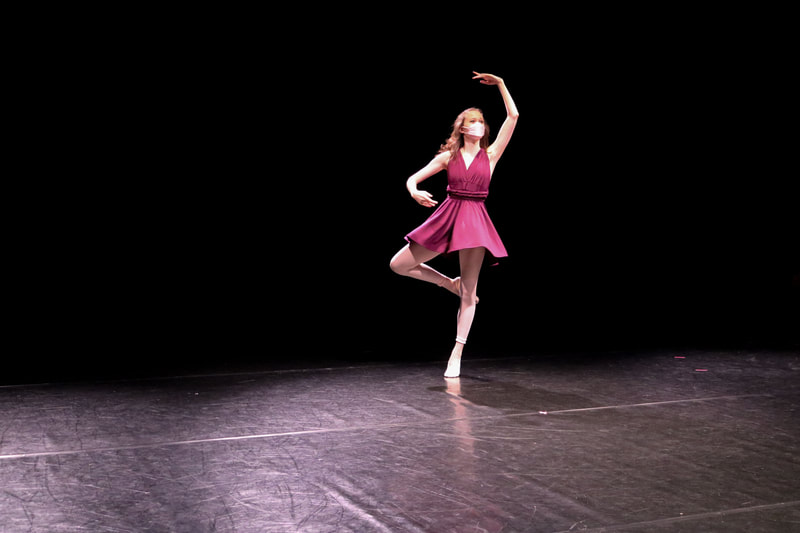 Dancer with pink mask and purple dress doing a passé on stage.