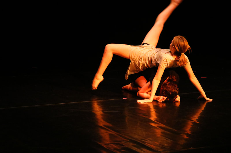 Dancer with gray dress doing a roll with pointed feet on top of a dancer in a black dress.